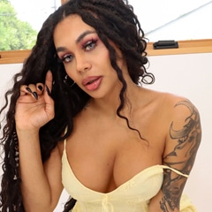 Latina trans model with tattoos kneeling on all fours on a bed with a red blanket